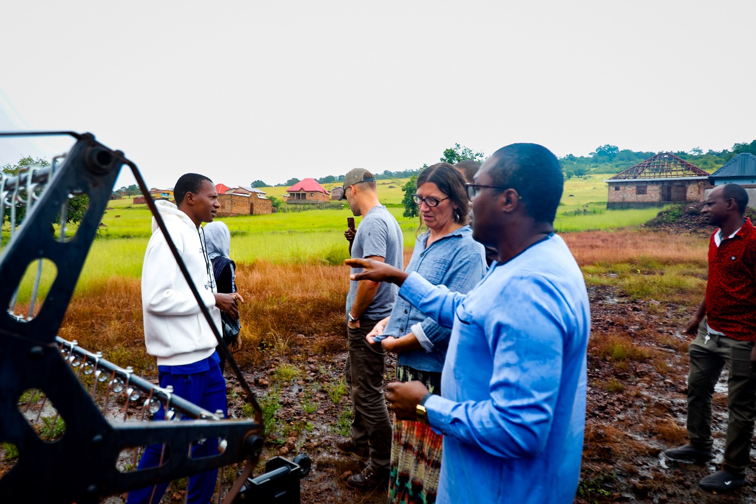 A man in blue traditional African clothing speaks with a woman next to farm equipment.