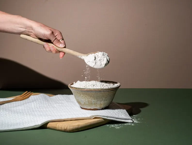 A hand holds a wooden spoon that is pouring fonio flour into a bowl below it.