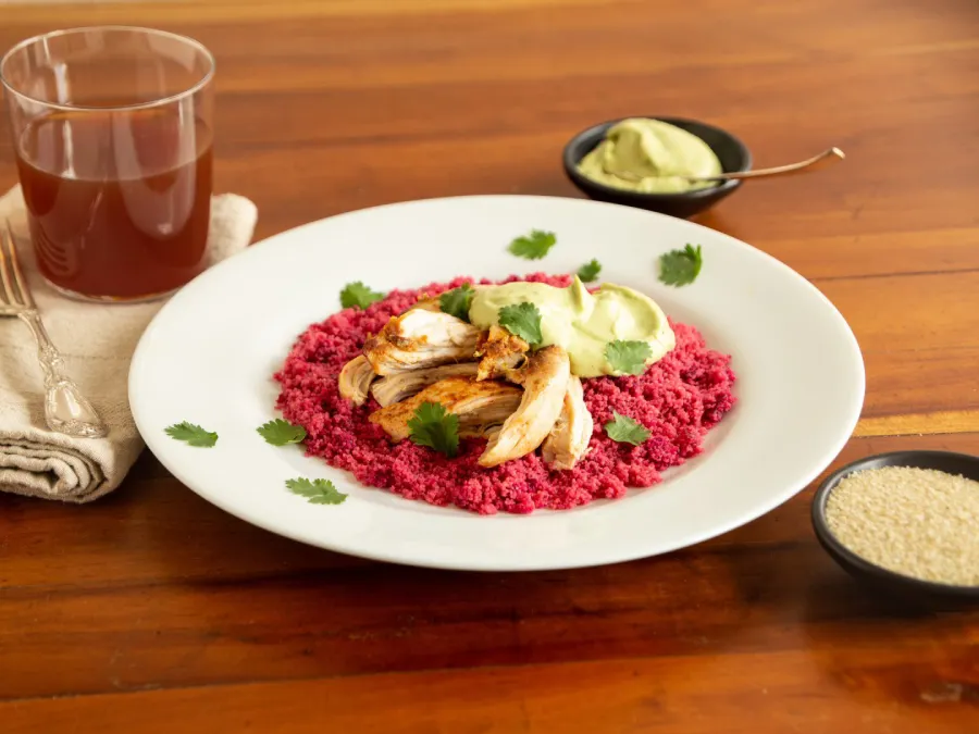 A plate of feet red fonio with chicken and avocado crema is displayed on a wood table with a glass of tea.